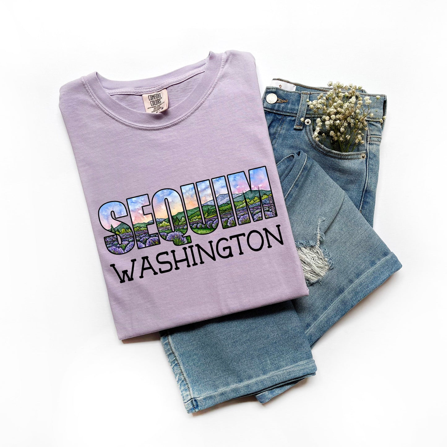 Folded purple t-shirt with 'Sequim Washington' text and graphic, paired with blue jeans on a white background.