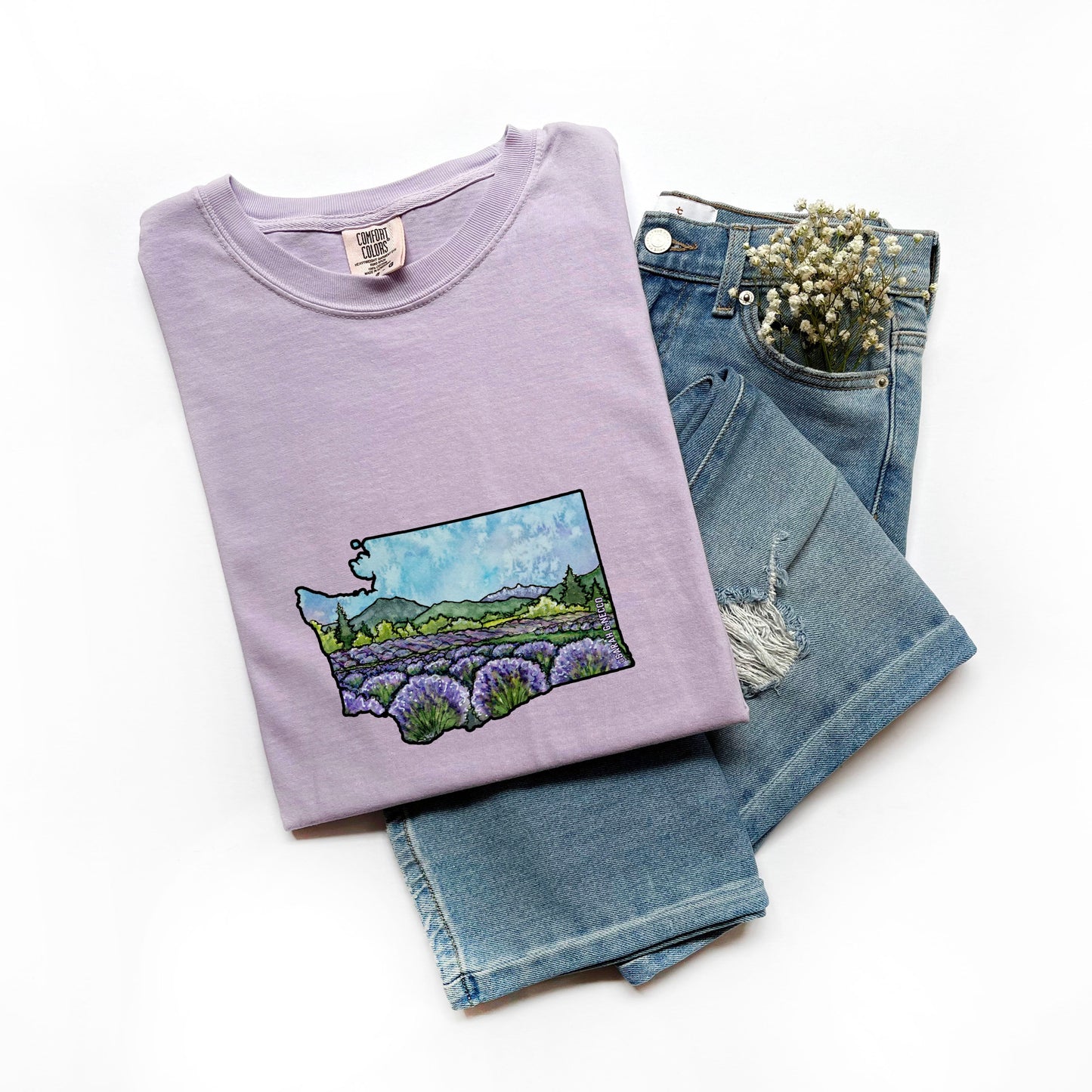 Folded lavender t-shirt with a watercolor WA State graphic design on jeans against a white background