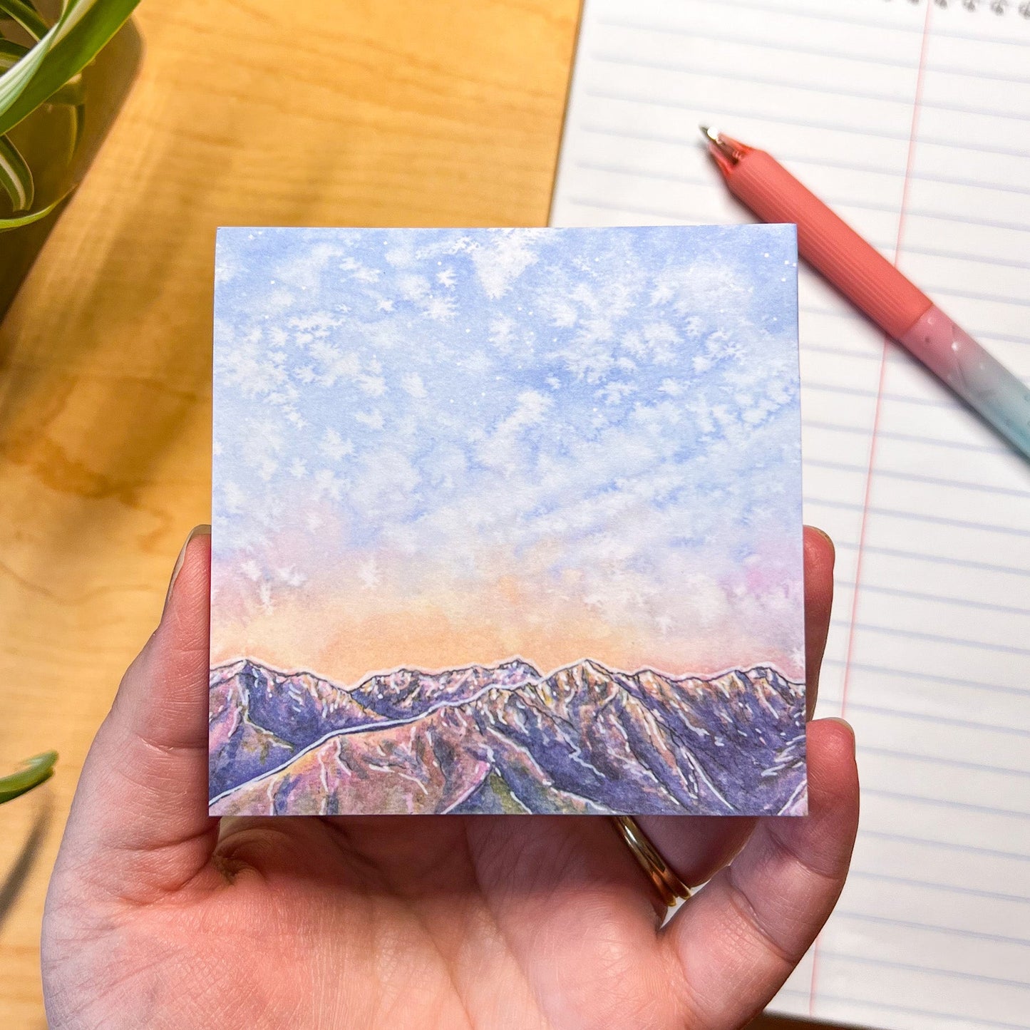 Hand holding a stick-note pad wit the design of mountains, with a pen and notebook in the background