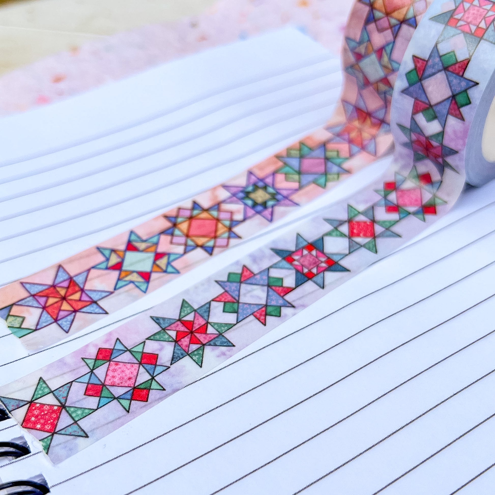 A close-up of a roll of patterned washi tape unrolled across lined notebook paper. The tape features a colorful geometric quilted star design in red, green, blue, yellow, and pink on a pale background.